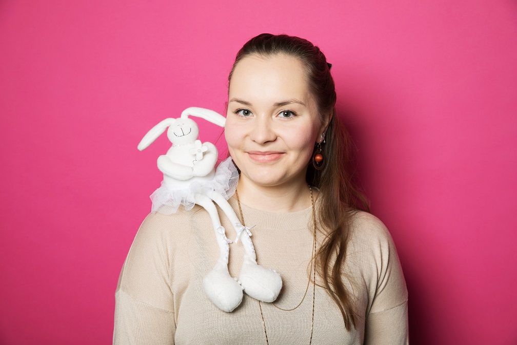 Pitkähiuksinen nainen, jolla vaaleanruskea paita. Olkapäällä pehmopupu. Pinkki tausta.
