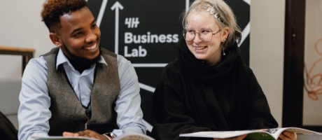 Kaksi nuorta istuu ja hymyilee. Käsissään vihkoset. Takana Business Lab -seinäke.