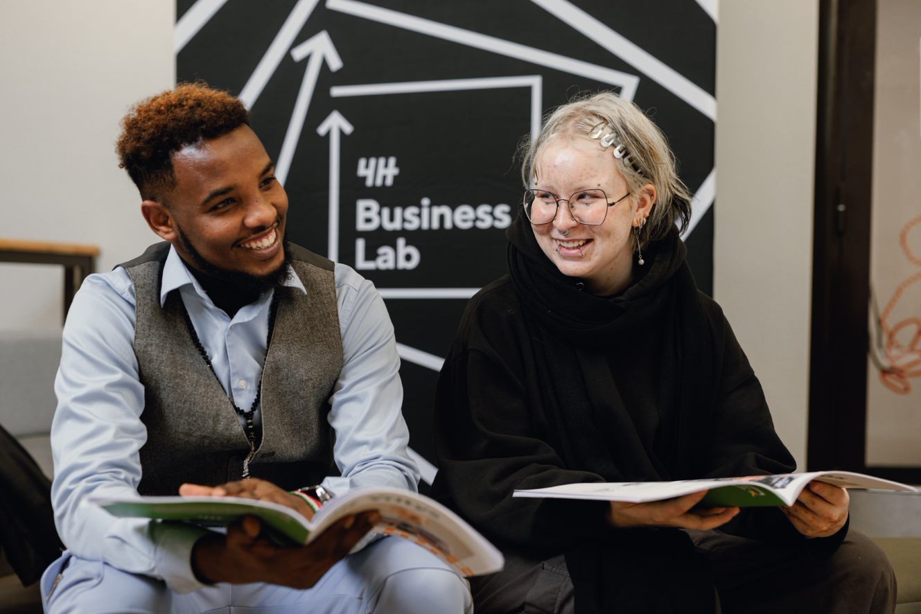 Kaksi nuorta istuu ja hymyilee. Käsissään vihkoset. Takana Business Lab -seinäke.
