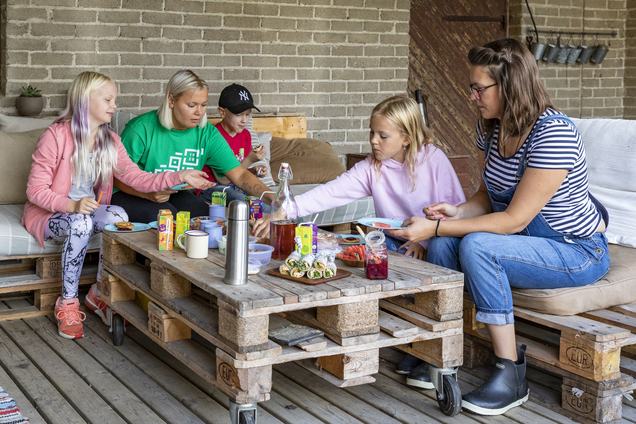 Vaihtolavoista tehdyn pöydän ja sohvan ääressä kolme kerholaista ja kaksi ohjaajaa syömässä välipalaa.