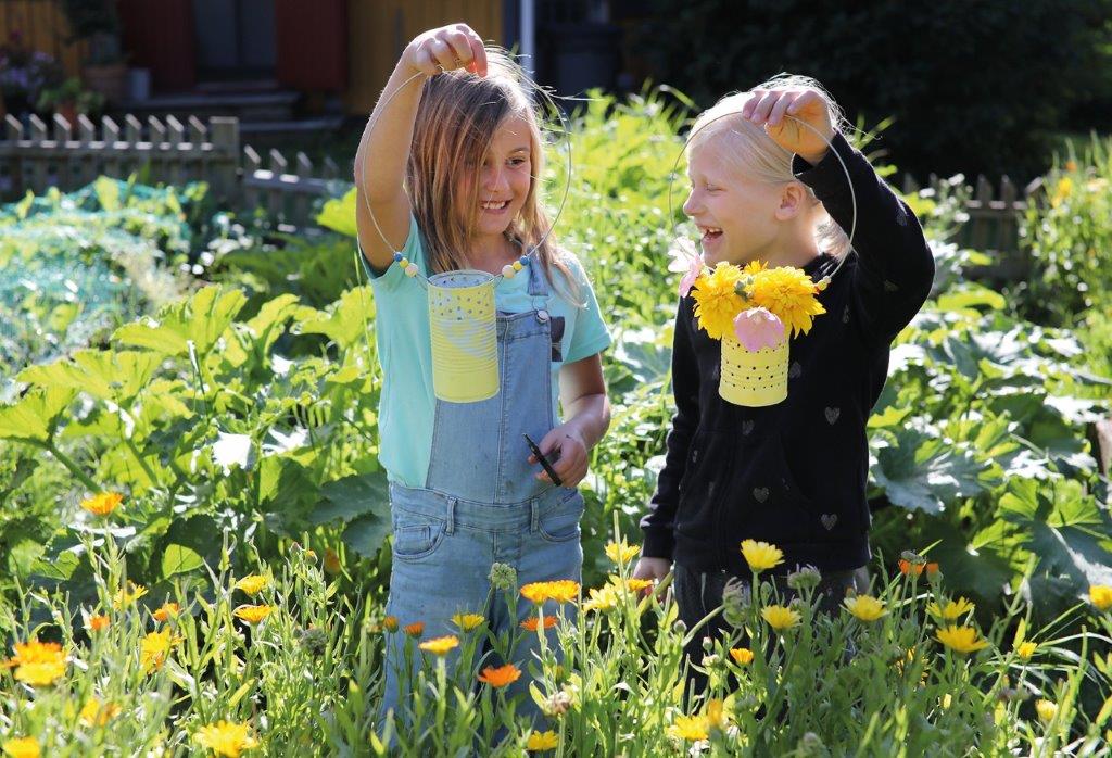 Kaksi lasta kukkakedolla. He hymyilevät ja pitävät käsissään metallitölkistä askarreltuja lyhtyjä