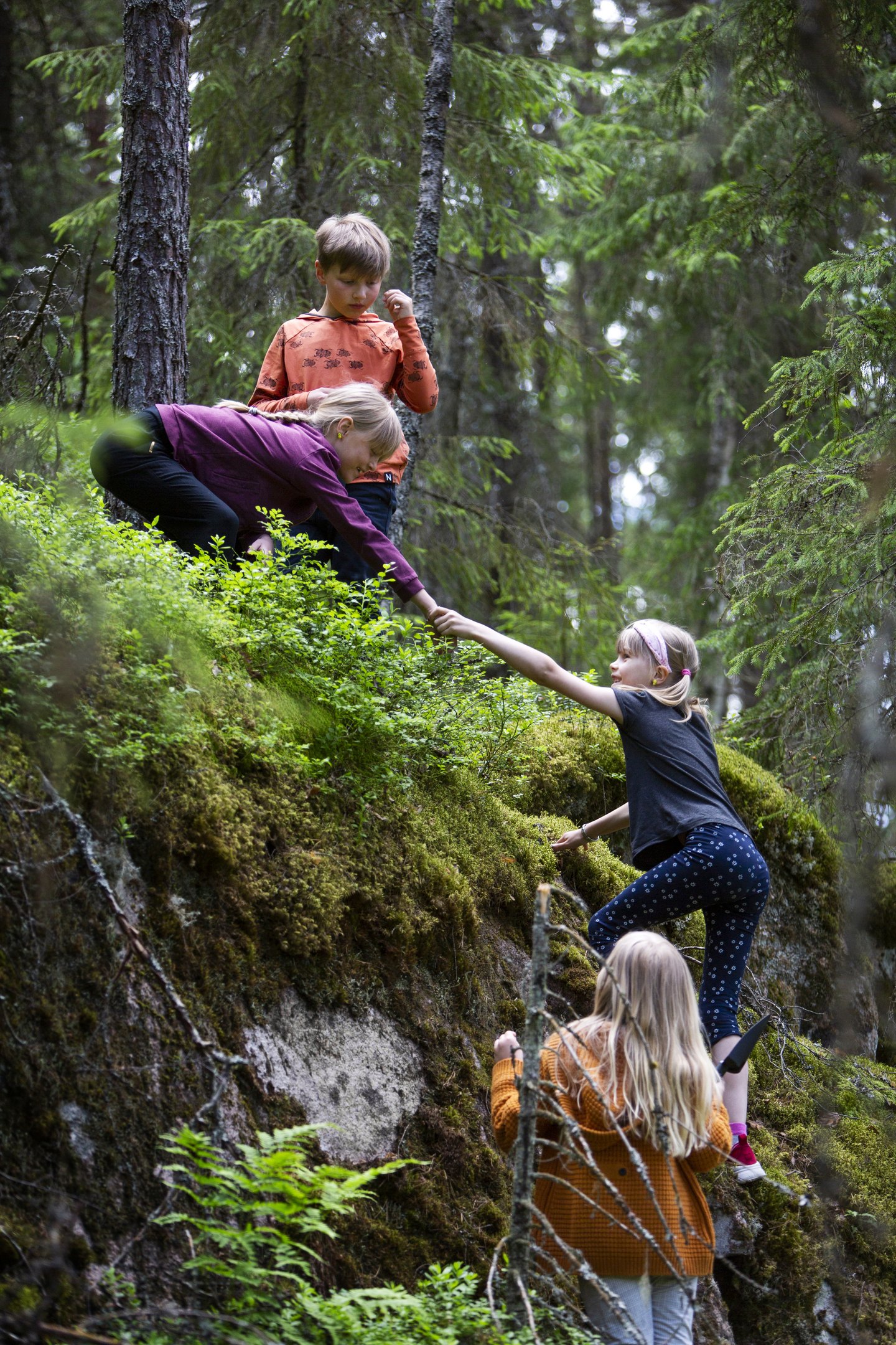 Neljä lasta kiipeämässä metsässä kalliolle, yksi auttaa toista kiipeämään ylös.
