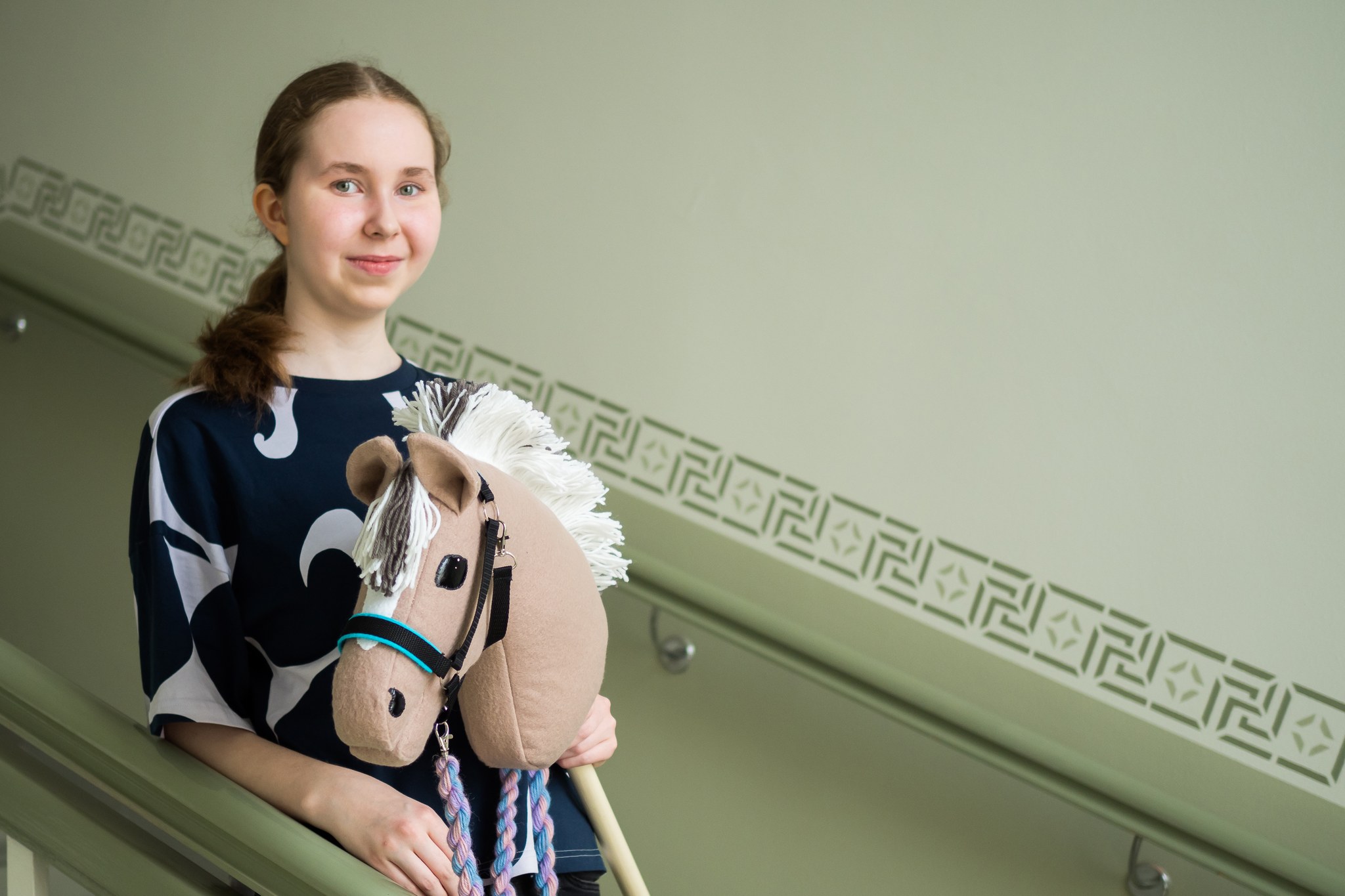 Tyttö hymyilee portaikossa itsetehdyn keppihevosen kanssa.