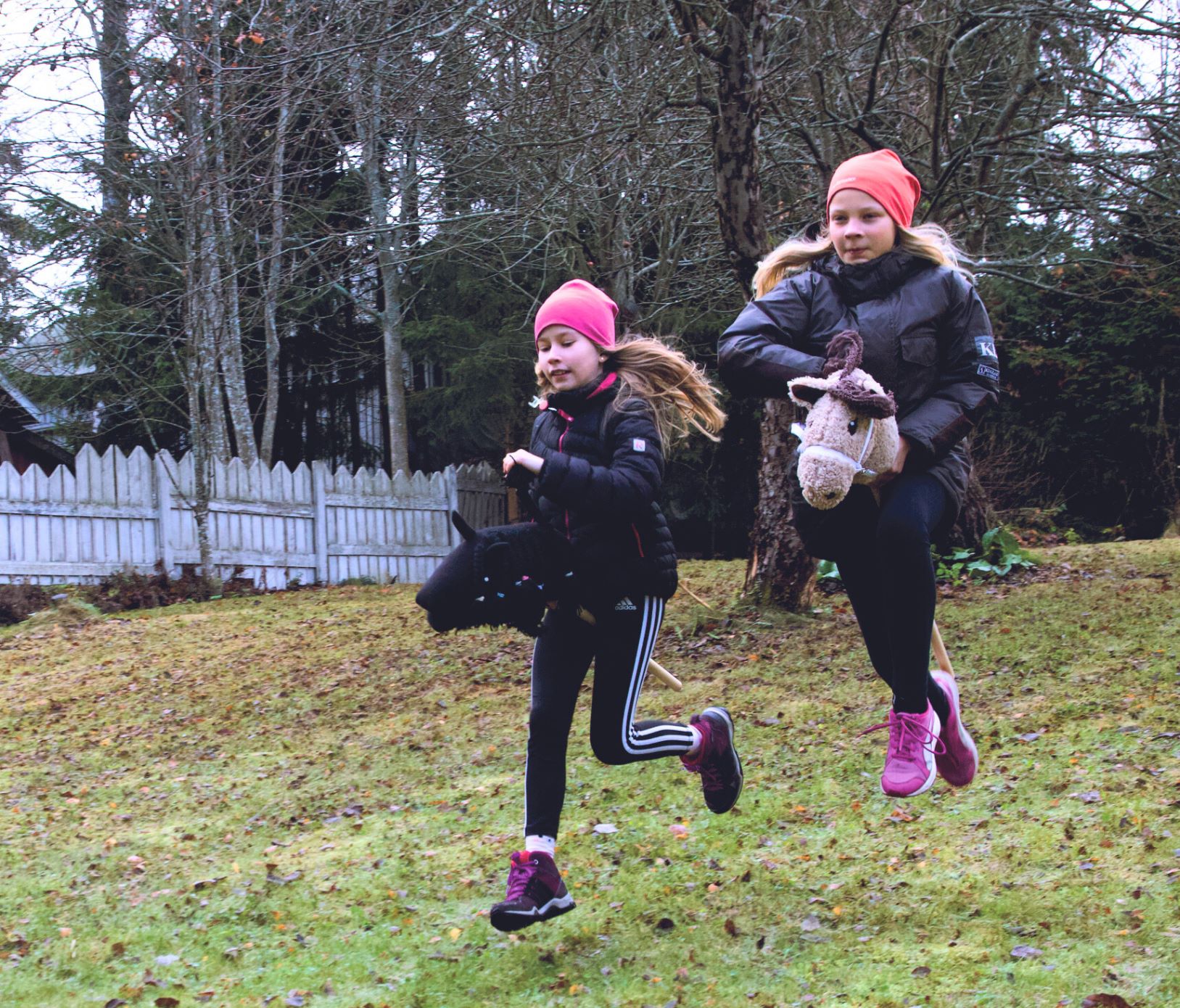 Kaksi lasta laukkaa keppihevosten selässä pihalla.